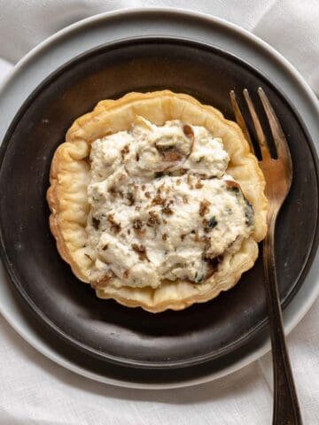tartlet with ricotta in grey plate and a light plat with a fork shown from above. The tartlet is showing the layer of ricotta and mushrooms and a sprinkle of truffle