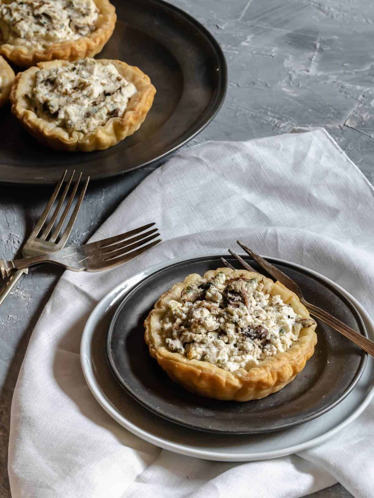 puff-pastry-tart-ricotta-and-mushrooms-truffle-on-plate-over-a-linen-with-fork-plate-with-three-other-tartlets-on the bottom-and-2-forks