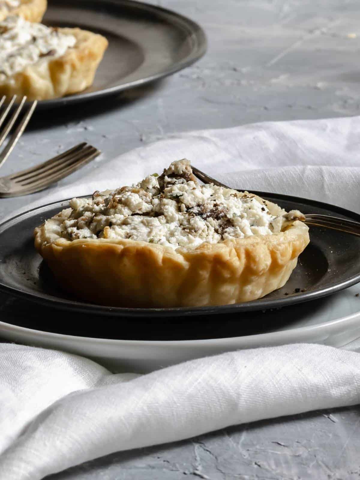 In-front-mini-puff-pastry-ricotta-and-mushrooms-truffle-on-a-plate-with-fork-on-the-bottom-plate-with-two-other-tartlets