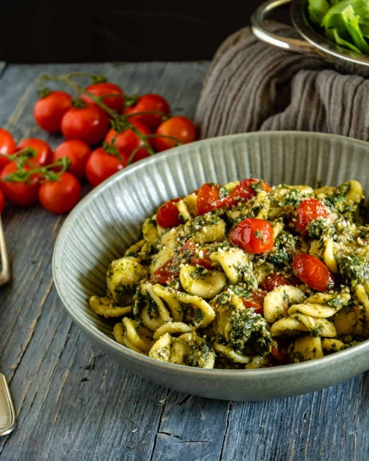 A view of Tuna and Spinach Orecchiette in a grey dish on a blue wooden table. The dish shown fresh cherry tomatoes and spinach on topy. On the side some tomatoes