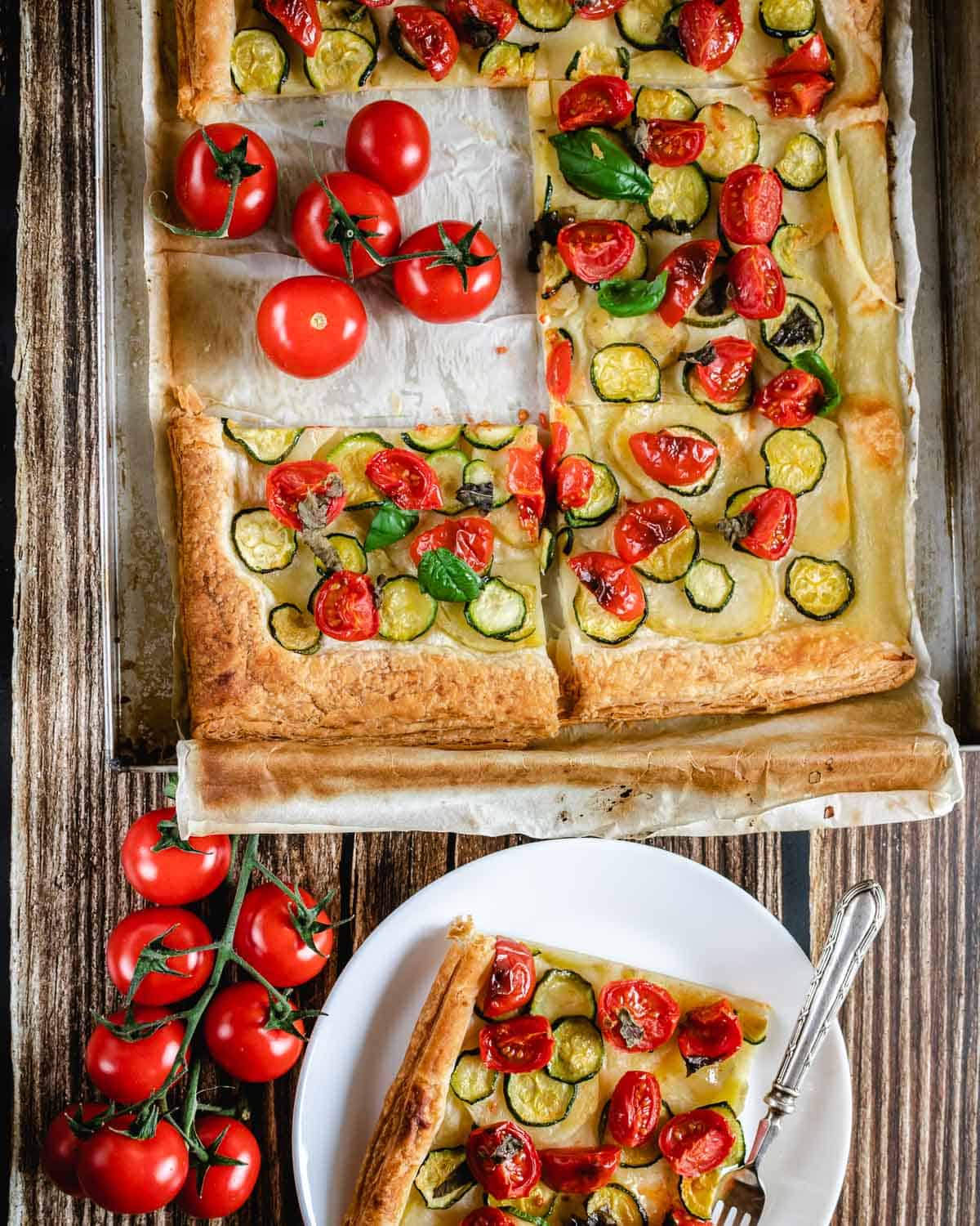 A view from above of a Zucchini Tart and a slice in a white plate with a fork on a wooden surface. The slice shown zucchini, potatoes and cherry tomatoes baked on top.