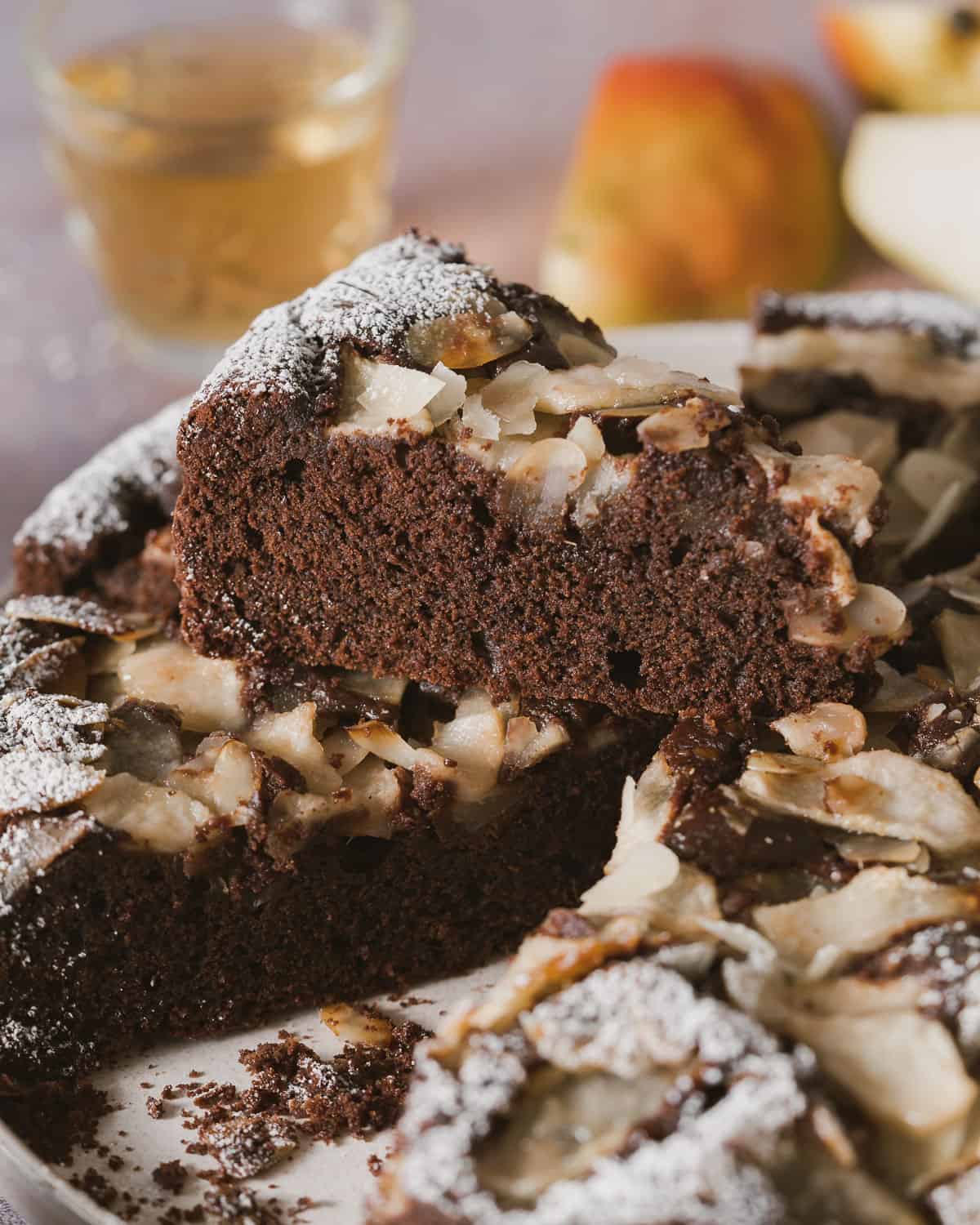A Chocolate and Pear Cake in a pan from above on a grey rustic table. The cake is sliced and a slice is out of the pan. It shown the pears baked inside and almonds on top