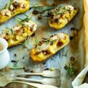4 half cut potato filled with mushrooms cheese and bacon on a baking tray with parchment paper. A white bowl with cheese cream and forks and spoon on the left