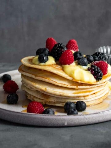 A straight on phot of a Pancakes stacks on a white plate and grey table. They are topped with custard and wild berries