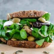 A Sandwich close up in a light wood board. The sandwich shown layers of eggplants, salad mozzarella and green olives