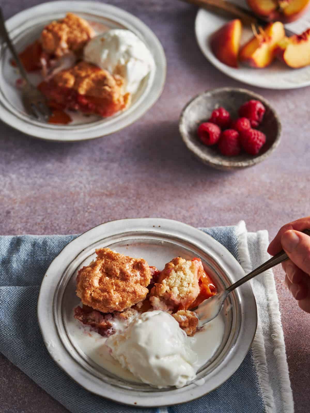 I'm cutting a slice of Peaches and Raspberries Cobbler in a white plate. The slice shown all fruit baked inside. It is served with some ice-cream