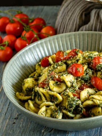 A view of Tuna and Spinach Orecchiette in a grey dish on a blue wooden table. The dish shown fresh cherry tomatoes and spinach on top. On the side some tomatoes