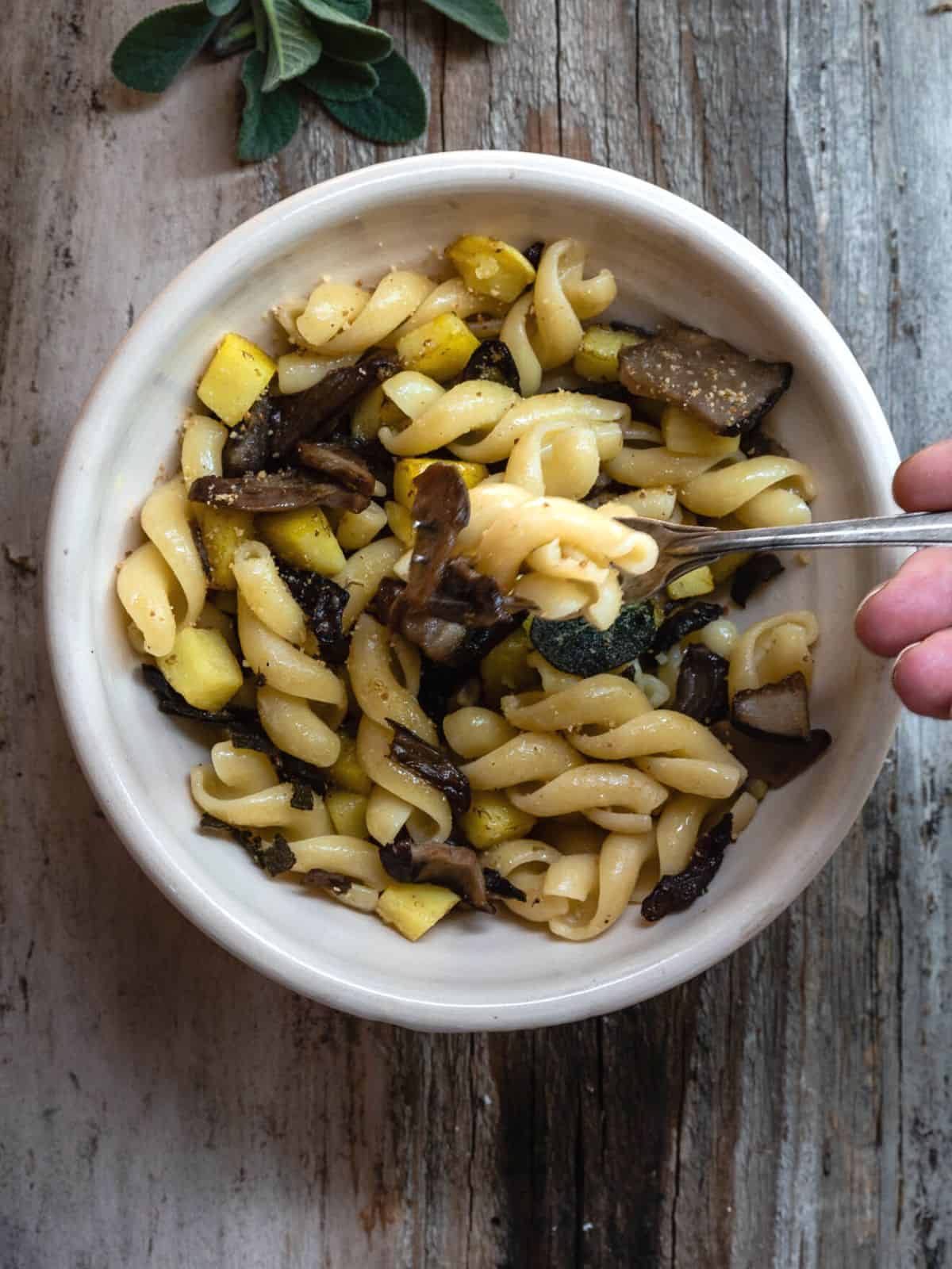 Me holding a fork with Mushrooms Pasta with Potatoes and Sage. It's in a light bowl on a rustic wooden table. The dish shown mushrooms, potatoes and browned sage on top. On the side some fresh sage