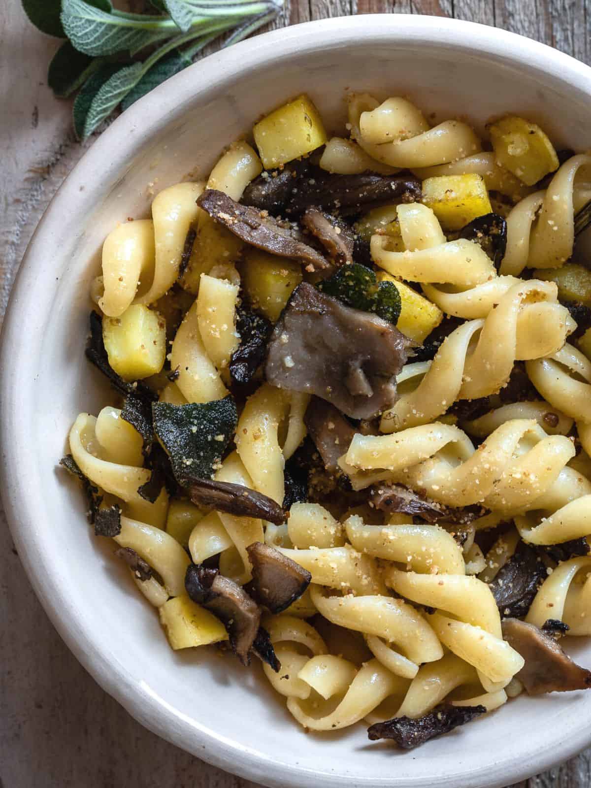 Mushrooms Pasta with Potatoes and Sage in a light bowl on a rustic wooden table. The dish shown mushrooms, potatoes and browned sage on top.
